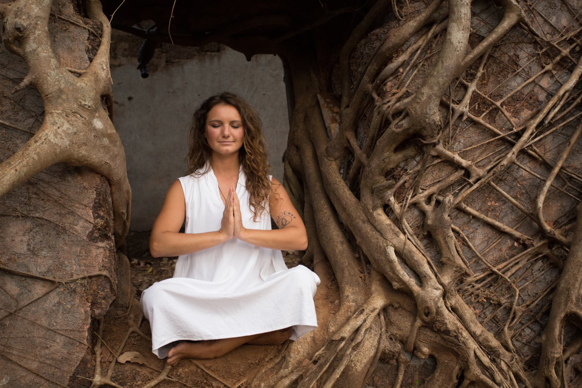 Izabella seated cross-legged with hands in prayer position in a stone alcove wrapped in tree roots